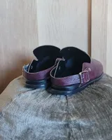 Zuecos de cuero vegano en color bordó con acabado texturizado. Presentan un diseño de punta cerrada con una tira ajustable con hebilla metálica que puede usarse en el tobillo o sobre el empeine.