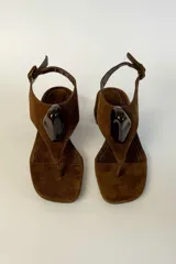 Sandalia de gamuza marrón con taco cuadrado bajo, diseño de tira tipo dedo y aplique decorativo negro brillante en el empeine. Cuenta con tira ajustable en el talón con hebilla.