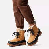 Bota de invierno Sorel modelo Tivoli V, confeccionada en gamuza impermeable color curry con refuerzos de goma negra en la base y el talón. Presenta interior forrado en piel sintética, cierre con cordones y suela de goma dentada color marrón.