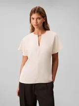 Blusa de manga corta Calvin Klein en color blanco marfil, confeccionada en tejido de crepé de chine. Presenta un diseño de corte regular con cuello en V adornado con un detalle de barra metálica horizontal.