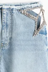 Jean de pierna recta en denim de algodón rígido, con diseño de aberturas laterales en la cintura decoradas con cristales brillantes. Presenta un corte holgado, cintura con borde desflecado y bajo deshilachado.