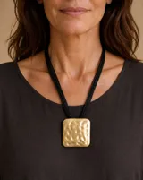 Collar de diseño geométrico con un gran dije rectangular de metal dorado con textura martillada y cordones negros múltiples.