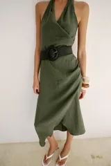 Vestido midi verde militar de cuello halter con escote en V cruzado y espalda descubierta. Cuenta con pliegues laterales, bajo asimétrico y un cinturón incorporado con hebilla redonda oscura.