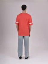 Remera de manga corta color rojo coral, con cuello redondo y detalles de franjas blancas en las mangas. Presenta un estampado frontal con texto que incluye "RHODE ISLAND", "1990" y "VARSITY LEAGUE".