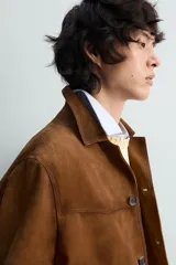 Cazadora de piel con acabado ante color marrón, corte relaxed fit, cuello bowling y manga larga con puño abotonado. Presenta bolsillos de vivo en la cadera y cierre frontal de botonadura.