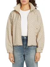 Campera deportiva Tommy Hilfiger color beige con estampado de logos en marrón. Tiene cuello alto, cierre de cremallera, dos bolsillos laterales con botones, puños elásticos y logo bordado en el puño.