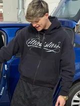 Campera con capucha negra, con cierre frontal completo y bolsillos estilo canguro. Presenta un estampado gráfico en blanco con letras cursivas en el pecho y diseño holgado.