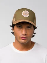 Gorro tipo trucker con frente de sarga en color verde oliva y visera curvada en color marrón. Presenta un parche circular bordado en el frente con el texto A Tractor Quality Company y paneles traseros de malla en color marrón con cierre ajustable snapback.