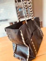 Bolso tipo tote de cuero negro con bolsillo externo y llavero interno. Tiene manijas cortas con diseño chevron y costuras blancas a contraste.