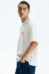 Remera de algodón blanco crudo, de corte oversized con hombros caídos y cuello redondo acanalado. Presenta pequeño estampado en el pecho con la inscripción "nas ©1994" en tono anaranjado.