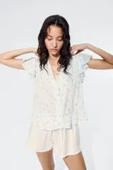 Blusa celeste claro con delicado estampado floretado azul, escote en V pronunciado, mangas cortas tipo mariposa y cierre frontal con botones. Presenta inserciones de encaje y alforzas decorativas en la parte delantera, además de un dobladillo con volados y detalles de encaje.
