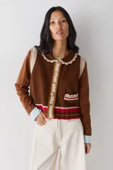 Chaleco de lana color marrón con cuello estilo bebé, cierre frontal con botones grandes y detalle de puntilla en cuello, sisas y bolsillo frontal. Presenta una franja decorativa en color rojo en el ruedo.