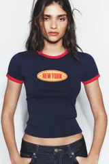 Camiseta de cuello redondo y manga corta, color azul marino con ribetes en color rojo en cuello y mangas. Presenta un estampado frontal ovalado con el texto New York en color naranja.