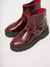 Botas tipo biker de caña baja color bordó, confeccionadas en cuero con plataforma de goma negra de 5 cm. Presentan detalles de costuras y bordados en hilo rojo en la parte delantera, elásticos laterales en el tobillo y una tira con hebilla metálica y tachas alrededor de la caña. Cuentan con cierre trasero para facilitar el calce.