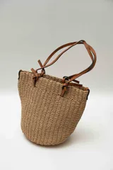 Bolso de mano tipo tote confeccionado en tejido texturizado efecto rafia en color beige, con asas dobles finas de cuero sintético marrón y detalles de nudos en los extremos.