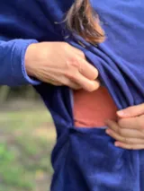 Polerón de plush azul con efecto batik en tonos rojizos, cuello alto holgado, mangas largas raglán, bolsillos frontales tipo parche y aberturas laterales para lactancia con dobladillo trasero más largo.