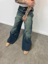 Jean de corte amplio con lavado desgastado en tono azul verdoso. Presenta bordados lineales en ambas piernas y cierres laterales metálicos que, al abrirse, revelan un panel de pana en color marrón. Cuenta con bolsillos amplios y botones personalizados.