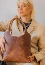 Matera de cuero color marrón con acabado desgastado, presenta costuras decorativas en los bordes superiores y manijas trenzadas. Incluye el logo de la marca Amadea grabado en el frente.