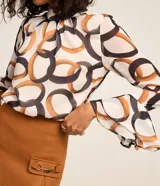 Blusa de chiffon con estampa de eslabones y argollas entrelazadas en tonos marrones y negros sobre fondo blanco. Presenta cuello alto, mangas largas acampanadas y cierre tipo gota en la espalda.