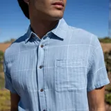 Camisa celeste con diseño a cuadros blancos, de manga corta, con cuello clásico, cierre frontal con botones marrones y bolsillo de parche en el pecho.