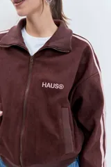 Cazadora de estilo deportivo en color marrón, confeccionada en tejido tipo gamuza. Presenta cierre frontal completo, cuello alto, puños y ruedo acanalados, y franjas laterales en tono rosado. Incluye el logo de la marca bordado en el pecho.