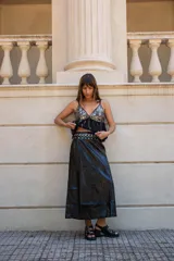 Pollera larga de ecocuero negro, de corte recto y tiro medio-alto. Presenta una cintura decorada con una hilera de ojales metálicos plateados.