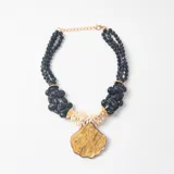 Collar de diseño artesanal compuesto por múltiples hileras de cuentas negras facetadas, secciones de cuentas negras irregulares y cuentas tubulares de color crema. Presenta un dije central grande en forma de concha marina con acabado marmolado en tonos marrones y dorados.