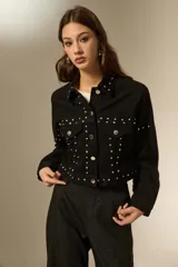 Chaqueta de jean negra con corte holgado, cuello camisero, cierre frontal con botones y detalle de strass y tachas metálicas distribuidas en el frente y mangas.