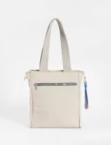 Cartera tipo shopper de color beige con diseño matelaseado de líneas onduladas y costuras en contraste azul. Cuenta con doble asa de hombro y un colgante decorativo de cordones multicolor en uno de sus laterales.