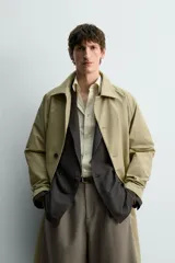 Trench de corte relajado confeccionado en tejido técnico italiano, color beige. Presenta cuello camisero, manga larga, bolsillos de vivo en la cadera y cierre frontal de botonadura simple con botones oscuros.