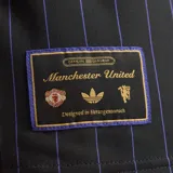 Camiseta de fútbol Adidas del Manchester United, modelo Terrace Icons. Diseño negro con sutiles rayas verticales moradas, franjas amarillas en los hombros y detalles bordados en dorado, incluyendo el trébol de Adidas y el escudo clásico del club.