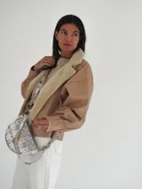 Bandolera de cuero color beige con correa ajustable y logo metálico dorado en el frente.