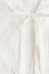Blusa blanca de algodón con escote en V, frente abierto con cierre de tiras finas para anudar, mangas largas y puños elásticos.
