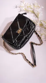 Bandolera negra acolchada con diseño de rombos, cierre dorado y correa de cadena dorada entrelazada con cuero sintético negro.