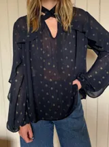 Blusa de gasa negra con estampado de lunares dorados y finas líneas verticales metalizadas. Presenta cuello con lazo, mangas largas con volados y puños elásticos, con un diseño de transparencia ligera.