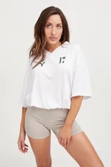 Camiseta blanca de poliéster microperforado con cuello en V y mangas cortas. Diseño oversize con cordón ajustable en la cintura y estampado minimalista con el número 17 y el logo de la marca en verde.
