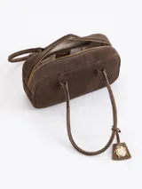 Cartera de mano de estructura rectangular, confeccionada en cuero descarne vacuno color verde oscuro. Presenta doble manija larga, cierre superior y un charm metálico dorado en forma de vaquita de San Antonio colgando de un tag lateral. Incluye el logo de la marca grabado en relieve en el frente.