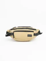 Riñonera color beige con detalles en negro, confeccionada en material resistente. Cuenta con dos compartimentos frontales con cierre, un bolsillo trasero oculto, manija superior y correa ajustable con broche de seguridad.