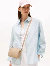 Cartera tipo camera bag de Tommy Hilfiger en color beige, con acabado granulado y logo de la marca en letras doradas en el frente. Cuenta con cierre de cremallera superior, compartimento principal, bolsillo interior abierto y bolsillo interior con cremallera. Incluye una correa ajustable de tejido textil con detalles distintivos de la marca.