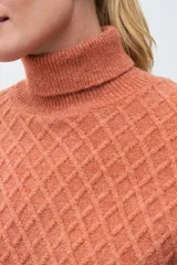 Polera de tejido de punto color ocre con cuello alto, diseño de rombos en relieve en el frente y mangas con detalle de trenzas laterales.