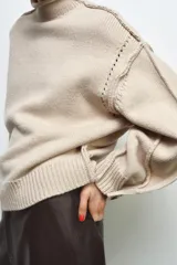 Jersey de punto color beige con cuello subido, manga larga y detalle de botones en los puños. Presenta costuras marcadas y acabados en rib.