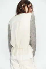Chaqueta de punto de cuello redondo y manga larga, con diseño combinado que incluye una capa interior en tono crudo. Presenta cierre frontal con botones y puños acanalados.