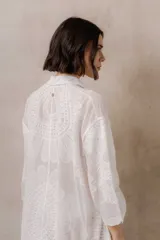 Camisola blanca de silueta amplia y caída liviana, con diseño clásico de cuello camisero, cierre frontal con botones y mangas tres cuartos, decorada con grandes motivos florales bordados.