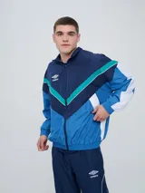 Campera deportiva Umbro modelo Beat, confeccionada en nylon ligero con diseño de bloques de color en blanco, azul marino y verde azulado. Presenta cierre frontal, cuello alto y elásticos en puños y cintura.