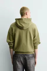Sudadera con capucha de corte relajado en color verde oliva con efecto lavado. Presenta un bolsillo frontal tipo canguro y un estampado gráfico en el pecho con letras en tonos amarillos y negros. Cuenta con puños y bajo acanalados.