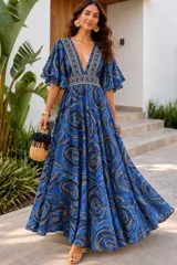 Vestido largo azul con estampado abstracto en tonos oscuros y detalles ornamentales en el escote en V y la cintura. Posee mangas amplias con volados y diseño fluido de gran movimiento.