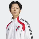 Campera deportiva Adidas de River Plate, color blanco, con detalles en rojo en los hombros y tres franjas negras en las mangas. Presenta cuello alto, cierre frontal y escudo del club bordado.