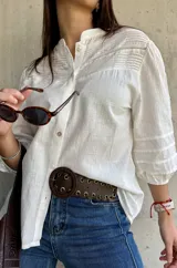 Camisa blanca de estilo romántico con cuello mao, botones frontales y mangas tres cuartos abullonadas con detalle de frunces. Presenta un diseño con textura sutil y costuras decorativas en la parte superior delantera.