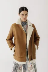 Campera tipo gamulán de cuero color camel con interior y solapas de corderito sintético en color crema. Presenta cierre frontal, bolsillos laterales y detalles de correas ajustables en los puños y la cintura.