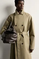 Trench regular fit de color beige, confeccionado en tejido técnico repelente al agua, con diseño de botonadura cruzada, cuello con solapas, mangas raglán con trabillas ajustables en los puños y cinturón del mismo tejido.